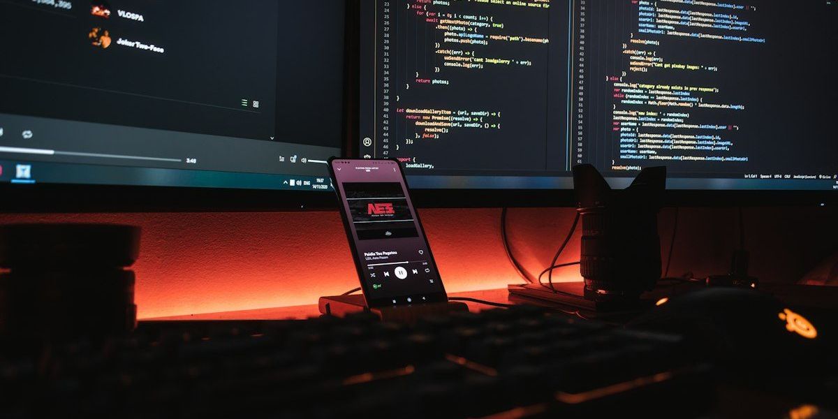 A smartphone displaying music on a desk with computer monitors showing code