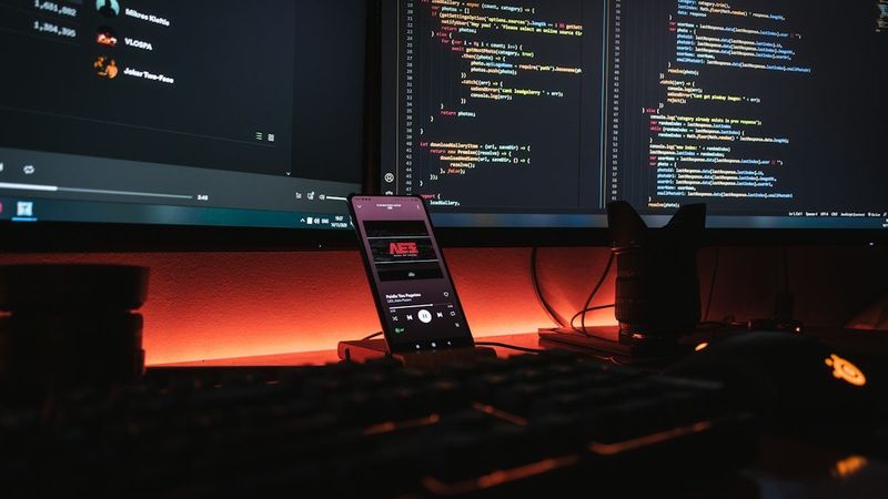 A smartphone displaying music on a desk with computer monitors showing code